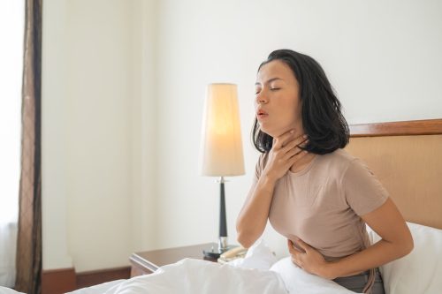 Mulher sentada na cama com a mão no pescoço e dor de garganta após cirurgia de amígdalas